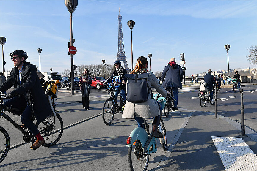 Radfahrende in Paris Menschen auf Fahrrädern überqueren eine Straße in Paris. Im Hintergrund ist der Eiffelturm zu sehen.
